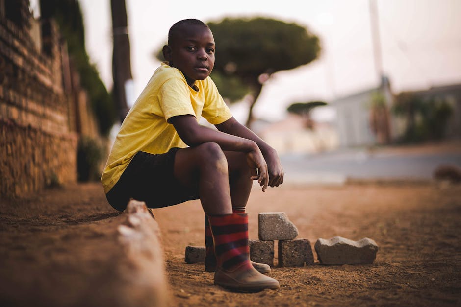A young boy in a yellow shirt sits outdoors on a quiet street during summer.